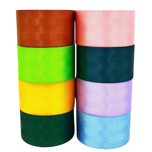 Puede incluir: Apilados rollos de cincha de nailon de colores. Los colores incluyen marrón, verde, amarillo, verde azulado, rosa, lavanda y azul claro. Los rollos, bien enrollados, muestran una textura tejida.