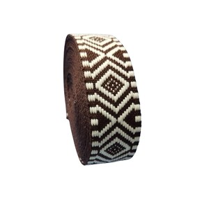 May include: A roll of brown and white patterned ribbon. The ribbon features a repeating geometric design of diamonds and zigzags in a contrasting white color. The ribbon is tightly wound, showcasing the detailed weave.