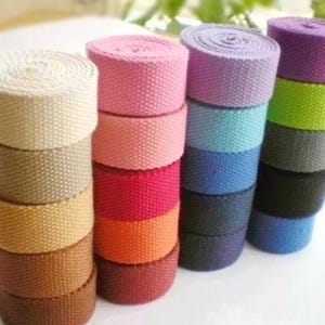 Puede incluir: Apilados rollos de cincha de lona en tonos beige, rosa, rojo, naranja, morado, azul, verde y negro. Los rollos están bien enrollados, mostrando la textura tejida de la tela. Ideal para manualidades y proyectos de bricolaje.