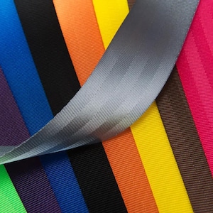 Peut inclure: Gros plan sur une variété d'échantillons de sangles de ceinture de sécurité colorées. Les couleurs comprennent des nuances de bleu, noir, orange, jaune, marron, rose et gris. La sangle grise est au premier plan, les autres couleurs sont disposées en rayures verticales.