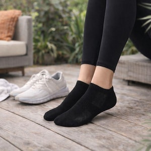 May include: A pair of black ankle socks are worn with black leggings. White athletic shoes and a white towel are on a wooden deck. A gray sofa with an orange pillow is in the background.