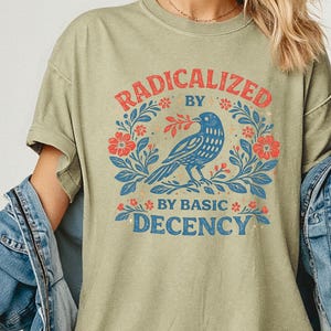 Op de afbeelding: Khaki T-shirt met de tekst "RADICALIZED BY BASIC DECENCY" in rood en blauw. Het ontwerp toont een blauwe vogel omringd door rode bloemen en groene bladeren. Het shirt heeft korte mouwen.