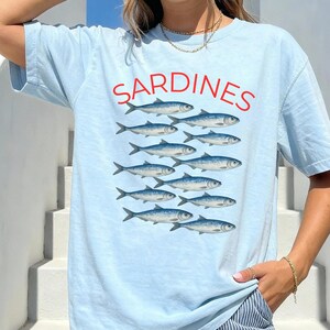 May include: Pale blue t-shirt featuring the word "SARDINES" in red, above a graphic of a school of fish. The fish are blue and white. The shirt is worn with blue and white striped trousers.