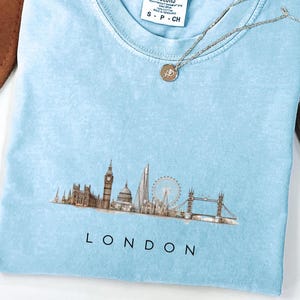 以下が含まれることがあります： ロンドンのスカイラインが描かれた水色のTシャツ。ビッグベン、ロンドンアイ、タワーブリッジなどのランドマークが特徴です。「LONDON」の文字が黒でプリントされています。シルバーのネックレスとペンダントがTシャツの上に置かれています。Comfort Colorsブランド。