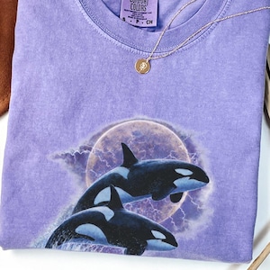 May include: A violet Comfort Colors t-shirt with a graphic of two orcas leaping in front of a full moon and lightning. A gold necklace with a small pendant is on the shirt. The shirt is folded and displayed on a white surface.