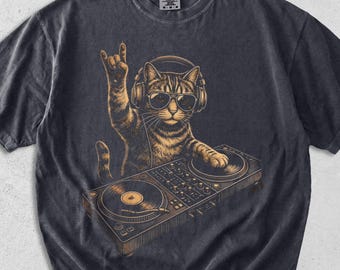 May include: Graphite-colored t-shirt featuring a cat DJ wearing headphones and sunglasses, with a hand gesture. The cat is positioned behind a turntable, with the text "Graphite" in the upper left corner.