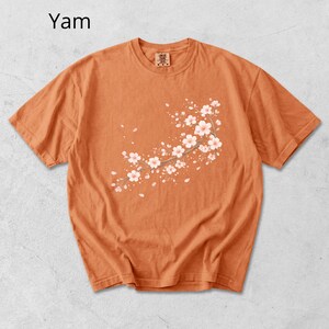 May include: Orange short-sleeve t-shirt with a floral branch design. The design features white flowers and light pink accents. The word "Yam" is printed in the upper left corner of the shirt.