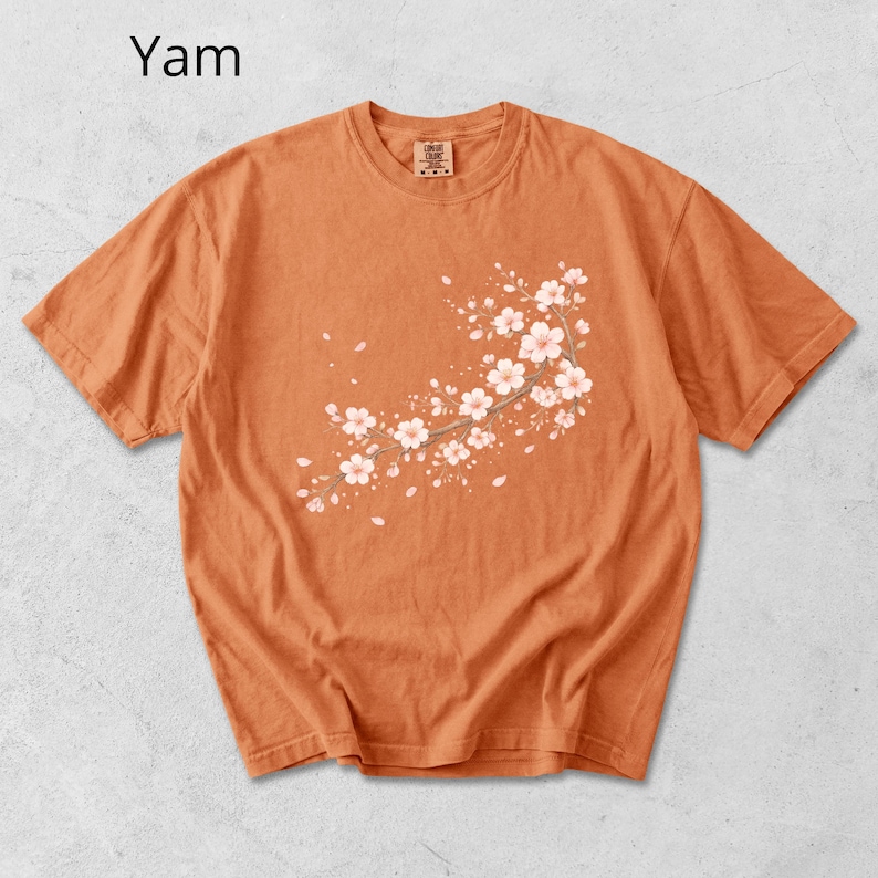 May include: Orange short-sleeve t-shirt with a floral branch design. The design features white flowers and light pink accents. The word "Yam" is printed in the upper left corner of the shirt.