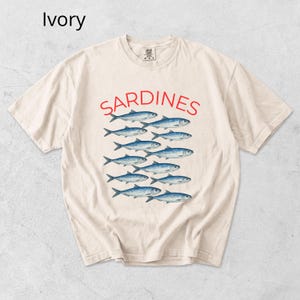May include: Ivory-coloured t-shirt with the word "SARDINES" in red above a graphic of blue fish. The shirt has a crew neck and short sleeves. The word "Ivory" is in the upper left corner.