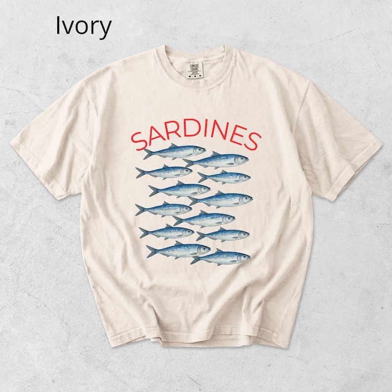 May include: Ivory-coloured t-shirt with the word "SARDINES" in red above a graphic of blue fish. The shirt has a crew neck and short sleeves. The word "Ivory" is in the upper left corner.