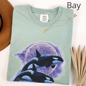 May include: A pale green Comfort Colors t-shirt with a graphic of two orcas jumping from the water against a purple moon and lightning background. The shirt has a crew neck and short sleeves. A gold necklace is draped across the shirt.