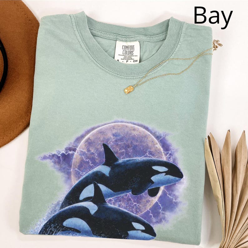 May include: A pale green Comfort Colors t-shirt with a graphic of two orcas jumping from the water against a purple moon and lightning background. The shirt has a crew neck and short sleeves. A gold necklace is draped across the shirt.