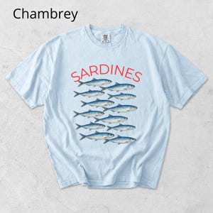 May include: Pale blue t-shirt featuring the word "SARDINES" in red above a graphic of fish. The word "Chambrey" is in the upper left corner. The shirt is a relaxed fit, suitable for everyday wear.