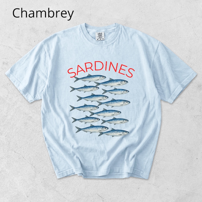 May include: Pale blue t-shirt featuring the word "SARDINES" in red above a graphic of fish. The word "Chambrey" is in the upper left corner. The shirt is a relaxed fit, suitable for everyday wear.