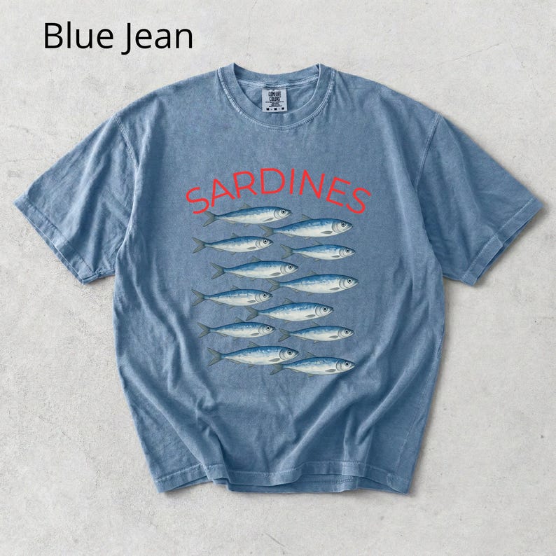 May include: Blue jean coloured t-shirt featuring the word "SARDINES" in red above a graphic of sardines. The shirt has a crew neck and short sleeves. The text "Blue Jean" is in the upper left corner.