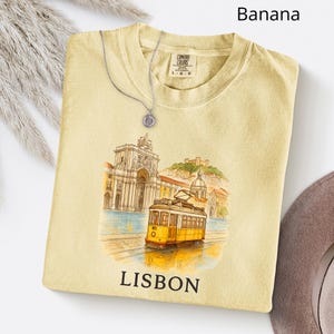 Puede incluir: Camiseta de color plátano con un gráfico de un tranvía amarillo en Lisboa, Portugal, con la palabra "LISBOA" debajo. Un collar plateado con un colgante está sobre la camiseta. La camiseta tiene una etiqueta Comfort Colors.
