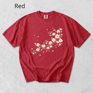 May include: Red t-shirt featuring a floral branch design with white and pink blossoms. The shirt has short sleeves and a crew neck. The word "Red" is printed in the upper left corner.