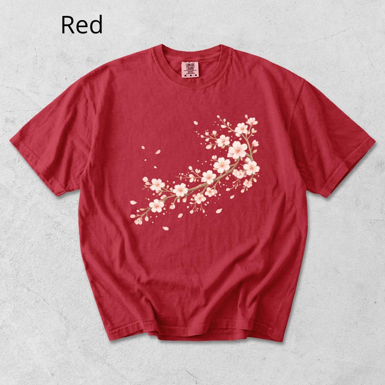 May include: Red t-shirt featuring a floral branch design with white and pink blossoms. The shirt has short sleeves and a crew neck. The word "Red" is printed in the upper left corner.