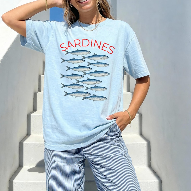 May include: Pale blue t-shirt featuring the word "SARDINES" in red, above a graphic of a school of fish. The fish are blue and white. The shirt is worn with blue and white striped trousers.