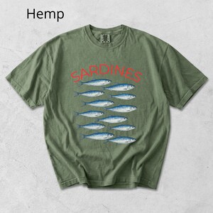 May include: Olive green t-shirt featuring the word "SARDINES" in red above a graphic of eleven blue and white fish. The word "Hemp" is printed in the upper left corner of the shirt.
