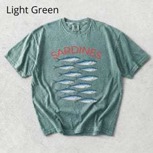 May include: Light green t-shirt featuring the word "SARDINES" in red above a row of blue fish. The shirt has a crew neck and short sleeves. The text "Light Green" is in the upper left corner.