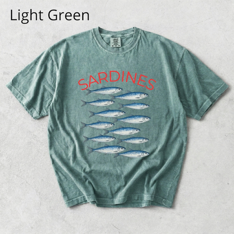 May include: Light green t-shirt featuring the word "SARDINES" in red above a row of blue fish. The shirt has a crew neck and short sleeves. The text "Light Green" is in the upper left corner.