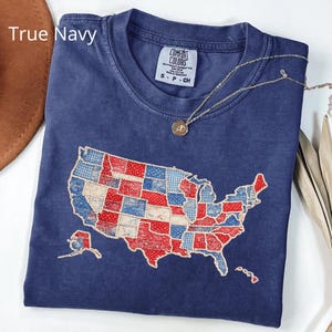 May include: A navy blue t-shirt with a graphic of the United States map, featuring a patchwork design in red, white, and blue. The shirt has the text "True Navy" and a Comfort Colors tag. A gold necklace with a pendant is draped on the shirt.