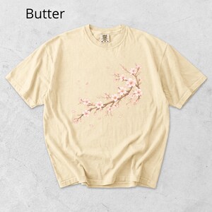 May include: Butter-colored t-shirt featuring a floral branch design with pink blossoms and falling petals. The text "Butter" is at the top left. The shirt has a crew neck and short sleeves.
