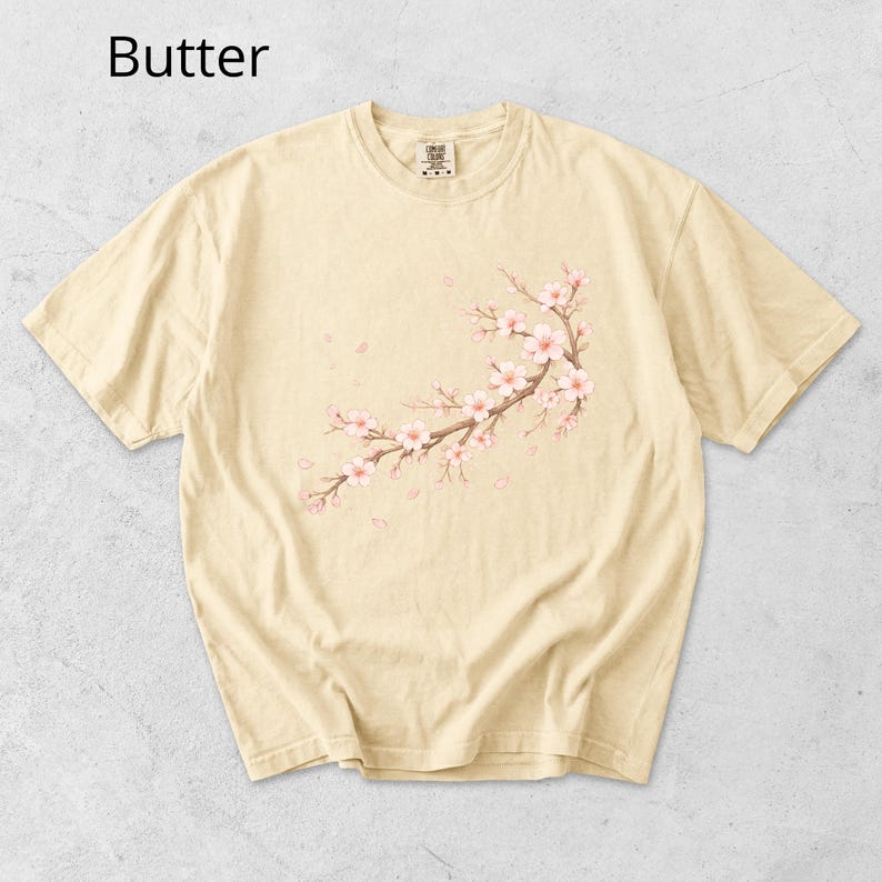 May include: Butter-colored t-shirt featuring a floral branch design with pink blossoms and falling petals. The text "Butter" is at the top left. The shirt has a crew neck and short sleeves.