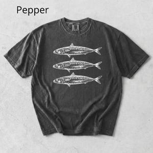 May include: Dark gray t-shirt with a crew neck featuring a white illustration of three fish. The word "Pepper" is printed in the upper left corner. The shirt has short sleeves and a slightly faded appearance.