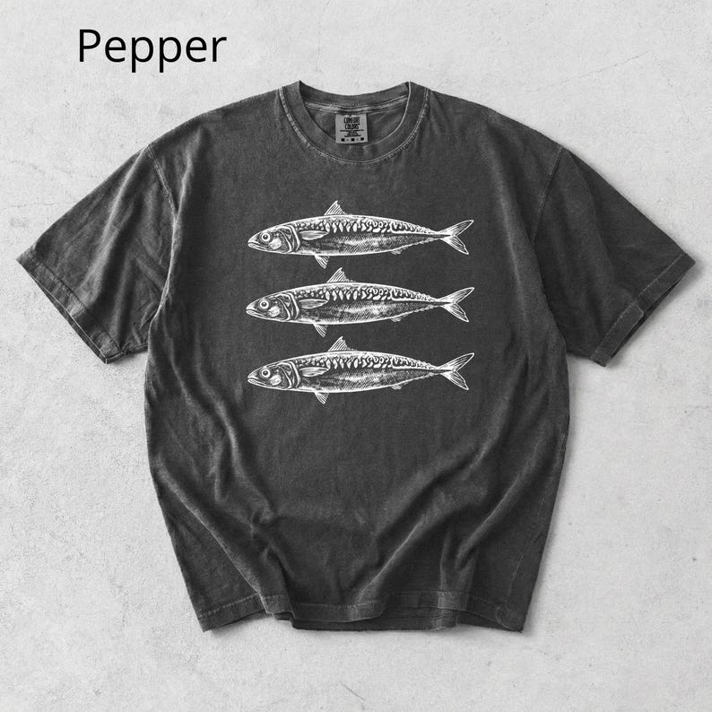 May include: Dark gray t-shirt with a crew neck featuring a white illustration of three fish. The word "Pepper" is printed in the upper left corner. The shirt has short sleeves and a slightly faded appearance.