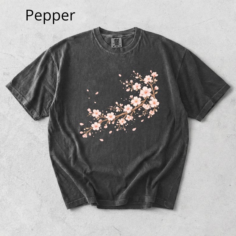 May include: Dark gray t-shirt with a floral branch design. The design features pink cherry blossoms and falling petals. The word "Pepper" is printed in the upper left corner of the image.
