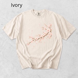 May include: Ivory-colored t-shirt featuring a watercolor illustration of a cherry blossom branch with pink flowers and falling petals. The shirt has a crew neck and short sleeves, perfect for casual wear.