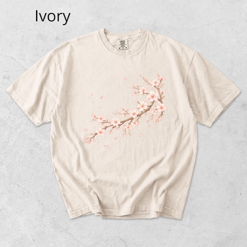 May include: Ivory-colored t-shirt featuring a watercolor illustration of a cherry blossom branch with pink flowers and falling petals. The shirt has a crew neck and short sleeves, perfect for casual wear.