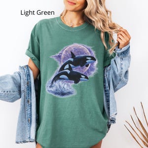 May include: Light green t-shirt with a graphic of two orcas leaping from ocean waves against a purple moon and cloudy background. The text "Light Green" is printed in the upper left corner of the image.