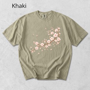 May include: Khaki-colored t-shirt featuring a floral branch design with pink blossoms and falling petals. The text "Khaki" is printed at the top left. The shirt has a crew neck and short sleeves.