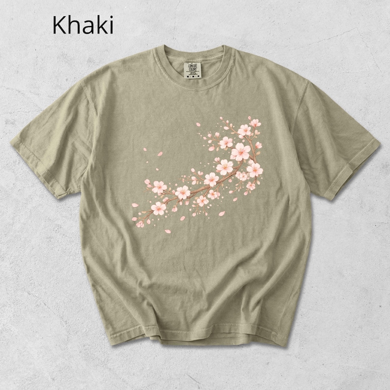 May include: Khaki-colored t-shirt featuring a floral branch design with pink blossoms and falling petals. The text "Khaki" is printed at the top left. The shirt has a crew neck and short sleeves.