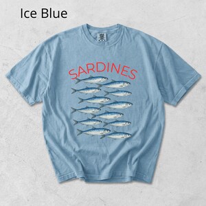 May include: Pale blue t-shirt featuring the word "Sardines" in red, above a graphic of fish. The shirt is a light blue colour.