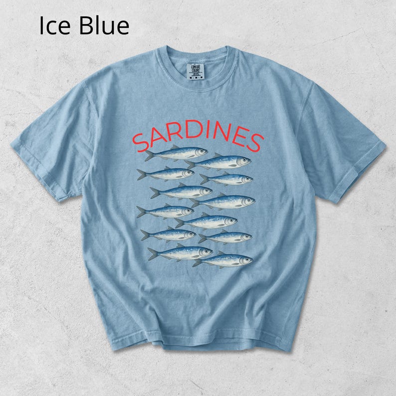 May include: Pale blue t-shirt featuring the word "Sardines" in red, above a graphic of fish. The shirt is a light blue colour.