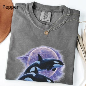 May include: A grey Comfort Colors t-shirt with a graphic of two orcas leaping in front of a purple moon and lightning. The shirt has a crew neck and short sleeves. A silver necklace with a small pendant is draped across the shirt.
