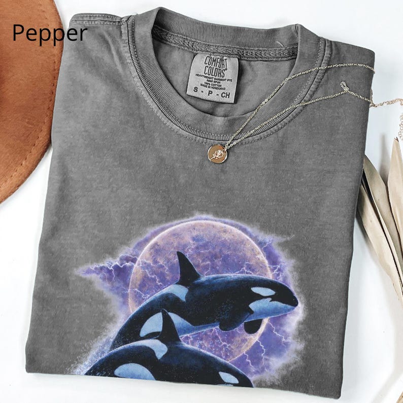 May include: A grey Comfort Colors t-shirt with a graphic of two orcas leaping in front of a purple moon and lightning. The shirt has a crew neck and short sleeves. A silver necklace with a small pendant is draped across the shirt.