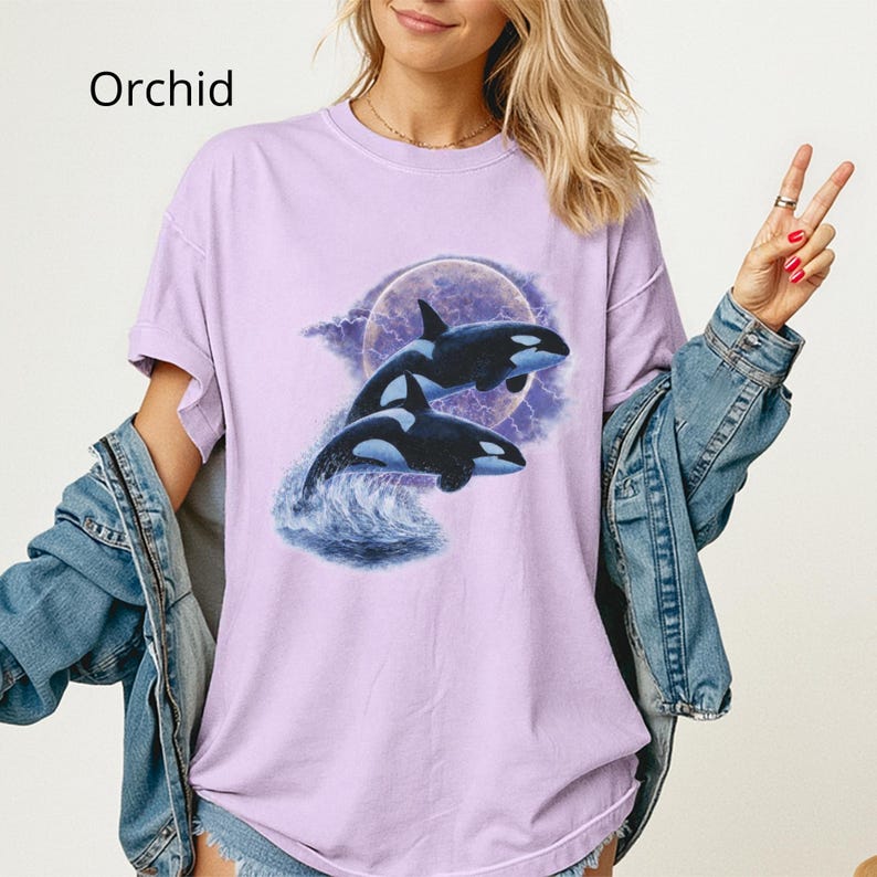 May include: Lilac t-shirt with a graphic of two killer whales jumping from the water under a full moon. The orcas are black and white, and the moon is set against a purple and white cloudy backdrop. The word "Orchid" is printed in white.