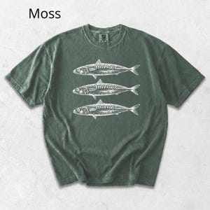 May include: Moss green t-shirt featuring a white illustration of three fish. The shirt has a classic crew neck and short sleeves. The word "Moss" is printed in the upper left corner. This casual tee is perfect for anyone who loves marine life.