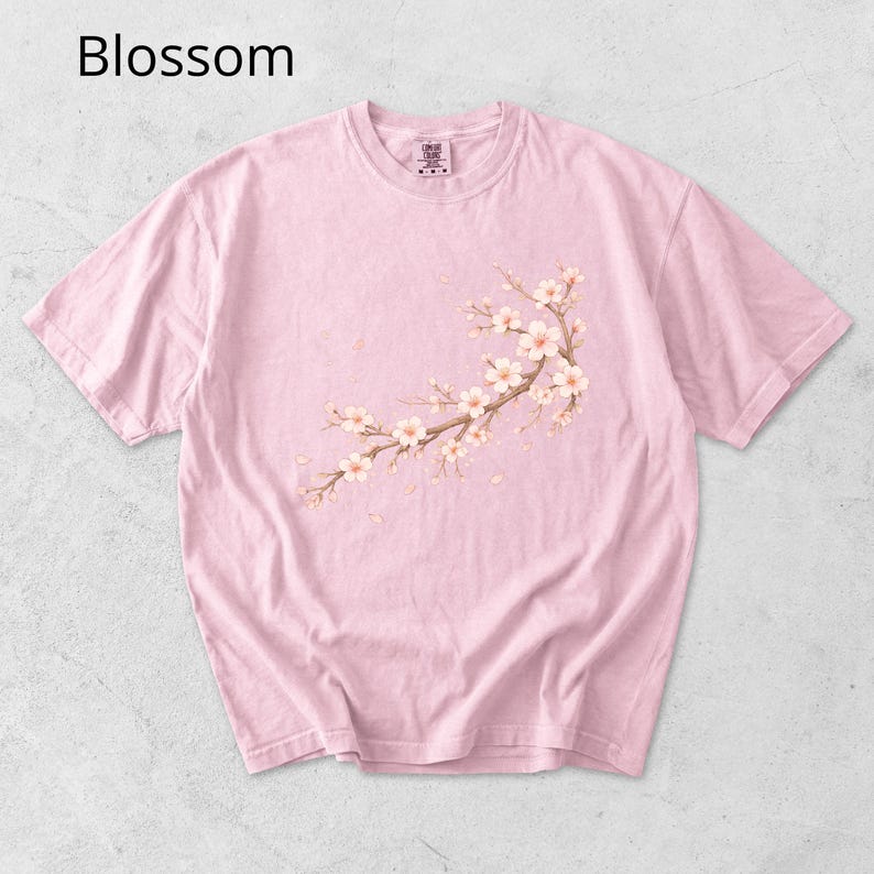 May include: A light pink t-shirt with a crew neck and short sleeves. The shirt features a floral branch design with white and pink blossoms. The word "Blossom" is printed in the upper left corner.