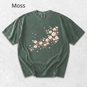 May include: Moss green t-shirt featuring a floral branch design with pink blossoms and falling petals. The short-sleeved shirt has a crew neck and a relaxed fit. The word "Moss" is printed in the upper left corner.