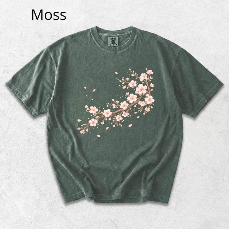 May include: Moss green t-shirt featuring a floral branch design with pink blossoms and falling petals. The short-sleeved shirt has a crew neck and a relaxed fit. The word "Moss" is printed in the upper left corner.