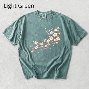 May include: Light green t-shirt featuring a floral branch design with pink blossoms and falling petals. The text "Light Green" is displayed at the top. This short-sleeved tee is a comfortable, casual wear option.