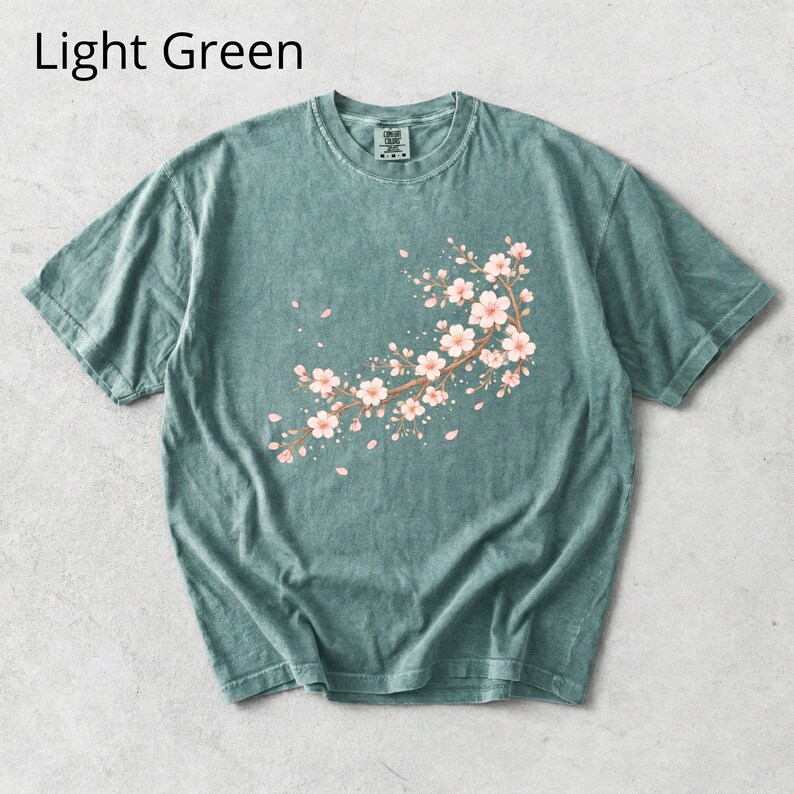 May include: Light green t-shirt featuring a floral branch design with pink blossoms and falling petals. The text "Light Green" is displayed at the top. This short-sleeved tee is a comfortable, casual wear option.