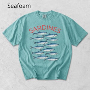 May include: Seafoam green t-shirt featuring the word "SARDINES" in red above a school of blue and silver fish illustrations. The t-shirt is short-sleeved with a crew neck design.