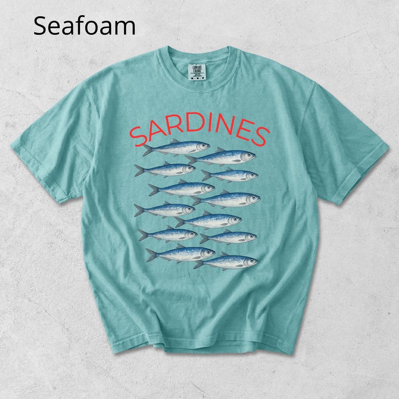 May include: Seafoam green t-shirt featuring the word "SARDINES" in red above a school of blue and silver fish illustrations. The t-shirt is short-sleeved with a crew neck design.